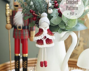 Puede incluir: Patrón de crochet para una pequeña muñeca de pelo blanco que lleva un vestido de crochet rojo y blanco y un gorro de crochet blanco. La muñeca está colgada de una jarra blanca con una planta verde y bayas rojas. El texto "Crochet Pattern - PDF" está en un círculo blanco en la imagen.