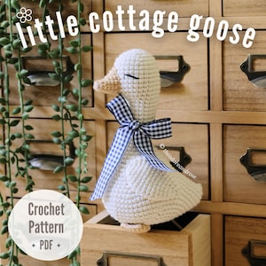 May include: A white crocheted goose toy with a tan beak and closed eyes, wearing a navy and white gingham ribbon around its neck. The text "little cottage goose" and "Crochet Pattern + PDF" are visible. The goose sits in a wooden drawer.