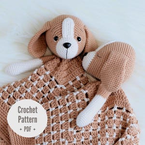 Puede incluir: Una manta de seguridad tejida a ganchillo en color canela y blanco con una cabeza de perro de ojos y nariz negros, unida a una manta cuadrada texturizada con patrón de rejilla. Una etiqueta blanca circular muestra Crochet Pattern PDF.