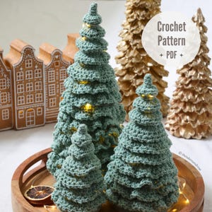 May include: Crocheted Christmas trees in varying sizes, displayed on a wooden tray. The trees are a soft green color and have a textured, layered design. The image also includes gold-colored decorative trees and gingerbread houses. The text "Crochet Pattern + PDF" is visible.