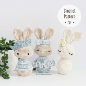 Op de afbeelding: Drie gehaakte amigurumi konijnen speelgoed. Het konijn links draagt een blauwe kroon, het konijn in het midden draagt een blauwe strik en het konijn rechts draagt een witte bloem.  Haakpatroon PDF.