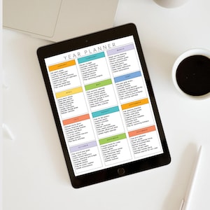 Może przedstawiać: Czarny tablet wyświetlający kolorowy roczny planer z sekcjami miesięcznymi. Planer zawiera listy dla każdego miesiąca, z nagł&oacute;wkami takimi jak "Styczeń" i "Marzec". Filiżanka kawy i długopis leżą na białej powierzchni.