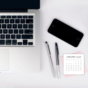 May include: A laptop keyboard, a black smartphone, two pens, and a small calendar for January 2026 on a white surface. The calendar is on a pink sticky note. The pens are silver and black.