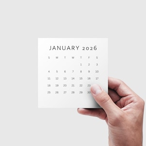 May include: A square white calendar page for January 2026 is held by a hand. The calendar displays the days of the month in a simple, minimalist design. The text "JANUARY 2026" is at the top.