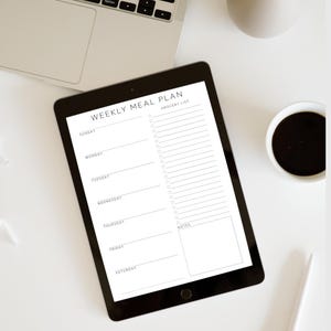 Może przedstawiać: Czarny tablet z napisem "WEEKLY MEAL PLAN" z sekcjami na każdy dzień tygodnia i listą zakup&oacute;w. W tle widoczny jest kubek kawy i laptop.
