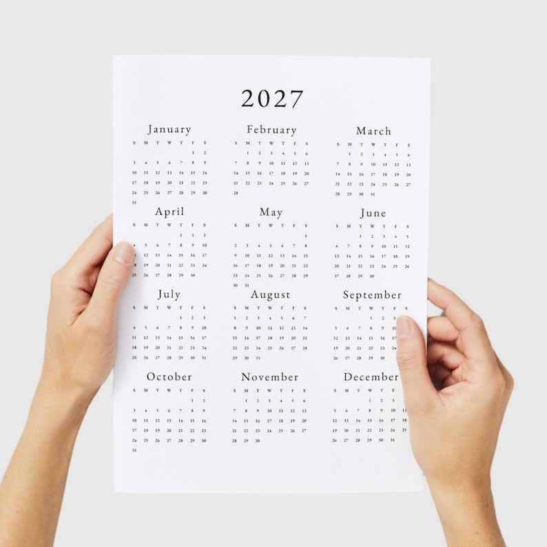 May include: A black and white calendar for the year 2027. Each month is displayed in a separate box with the days of the week labeled as S, M, T, W, T, F, S. The calendar is printed on white paper and is being held by a person's hands.