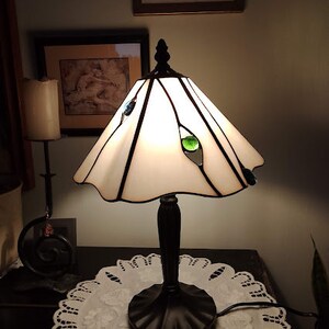 May include: A white stained glass table lamp with a black base and a white doily on a dark surface. The lamp has green and blue glass accents.