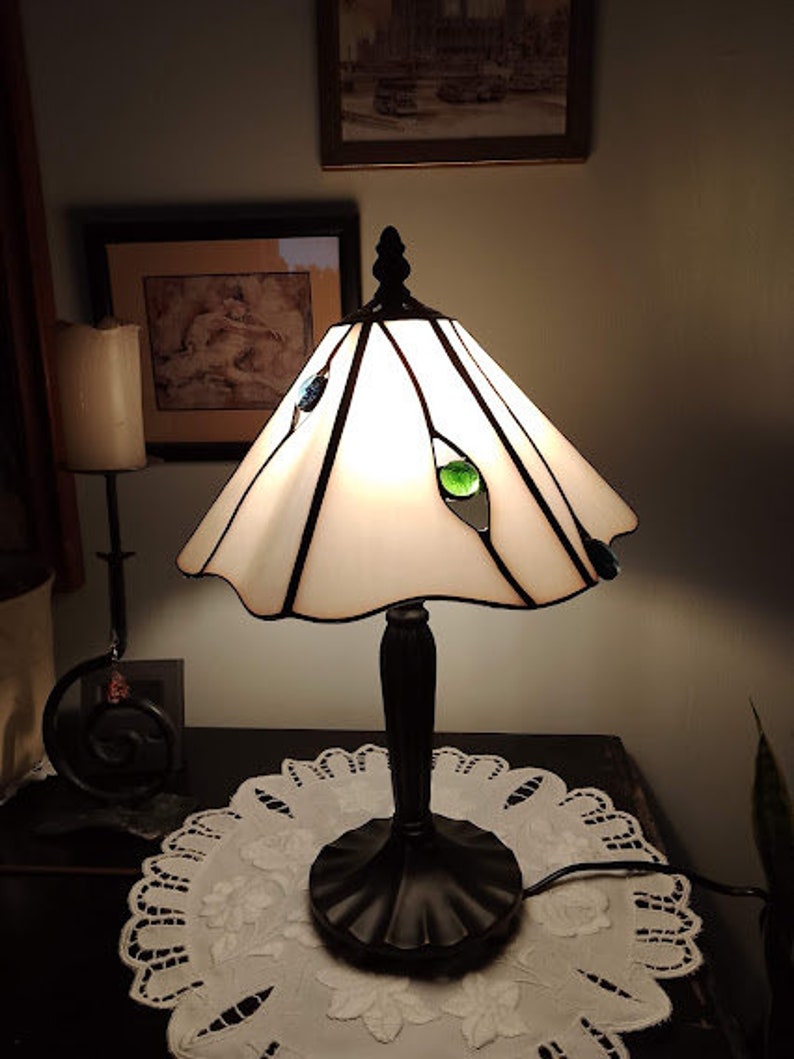 May include: A white stained glass table lamp with a black base and a white doily on a dark surface. The lamp has green and blue glass accents.