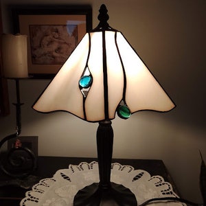 May include: A white stained glass table lamp with a black metal base and teal glass accents. The lamp is turned on and illuminating the room.