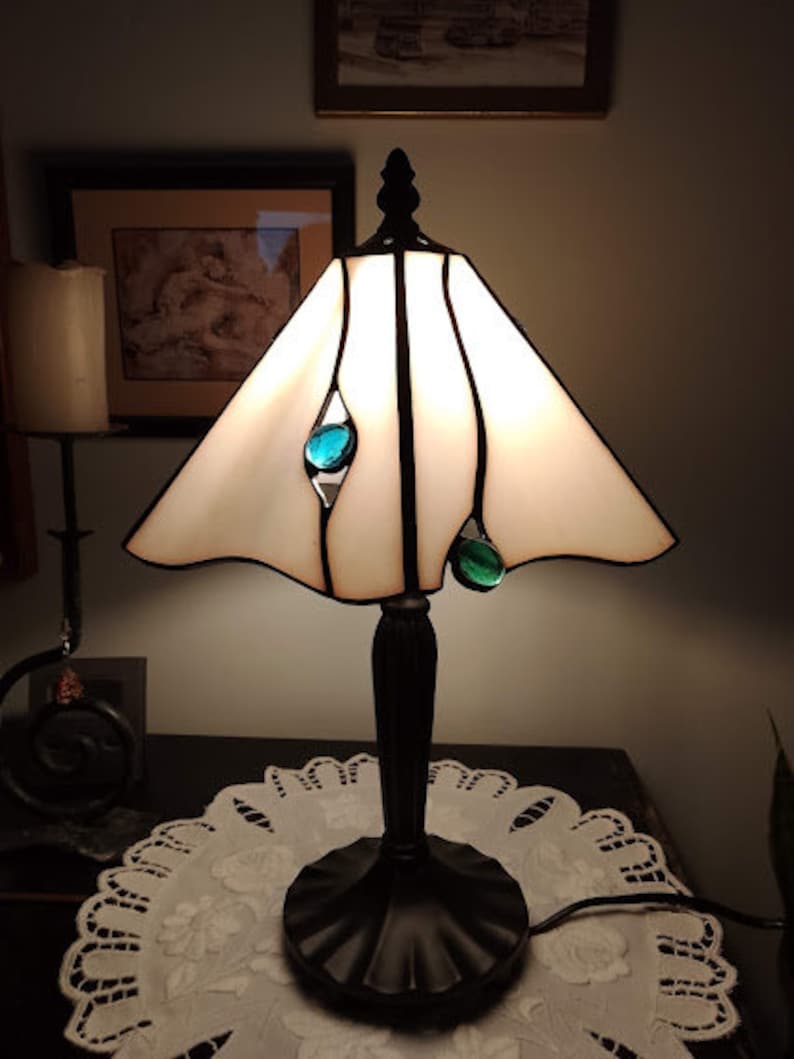 May include: A white stained glass table lamp with a black metal base and teal glass accents. The lamp is turned on and illuminating the room.