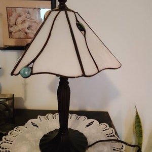May include: A white stained glass lamp with a black metal base and a white shade. The lamp has three small, round, blue and green glass accents on the shade.