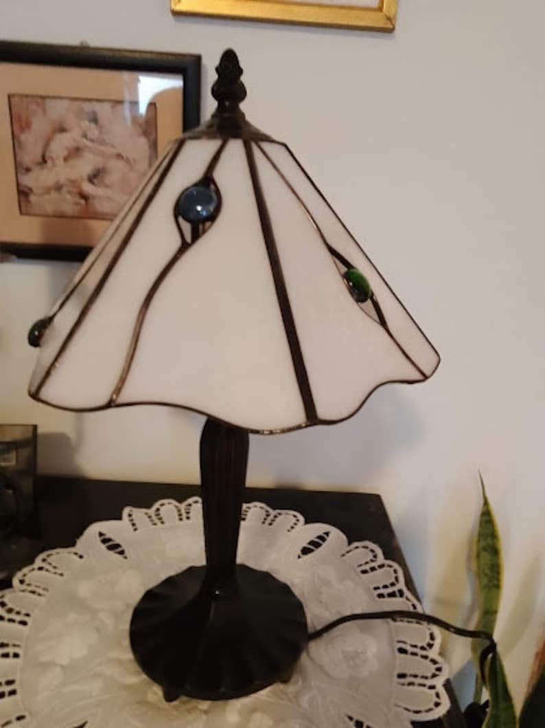 May include: A white stained glass lamp with a black metal base and three colored glass accents. The lamp is sitting on a white doily.
