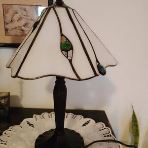May include: A white stained glass lamp with a black metal base and green glass accents. The lamp is sitting on a white doily on a dark surface.