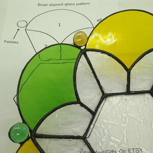 Op de afbeelding: Een glas-in-loodpatroon voor een kom, met een groen, geel en wit ontwerp. Het patroon is uitgelegd op een wit oppervlak met een diagram van het patroon en de tekst "Bowl stained glass pattern" bovenaan.