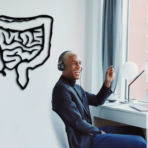 May include: A young man wearing headphones and a black blazer smiles while sitting at a desk with a laptop and a lamp. A black and white drawing of the human digestive system is on the wall behind him.