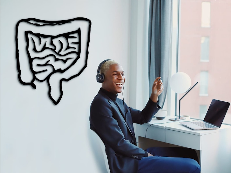 May include: A young man wearing headphones and a black blazer smiles while sitting at a desk with a laptop and a lamp. A black and white drawing of the human digestive system is on the wall behind him.