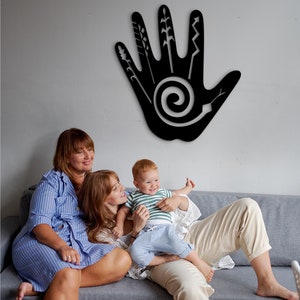 May include: Two adults and a child sit on a gray couch in front of a black wall decor. The decor is a handprint with a spiral design and various lines and shapes. The adults are wearing casual clothing and the child is wearing a striped shirt and blue jeans.