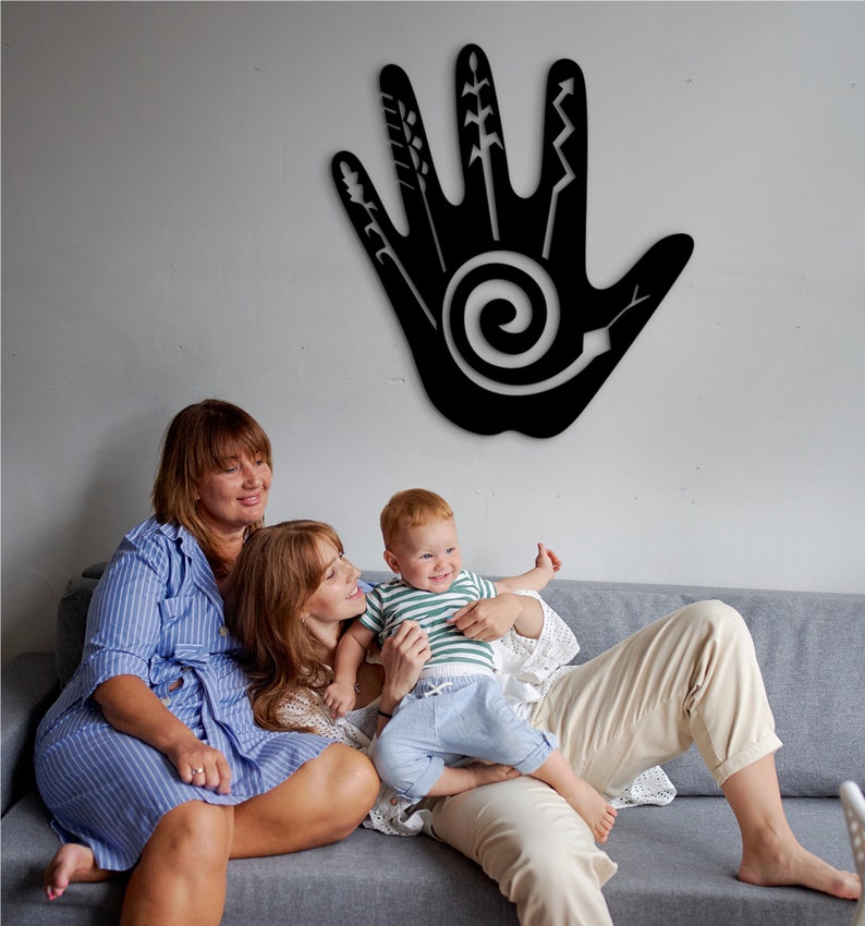 May include: Two adults and a child sit on a gray couch in front of a black wall decor. The decor is a handprint with a spiral design and various lines and shapes. The adults are wearing casual clothing and the child is wearing a striped shirt and blue jeans.