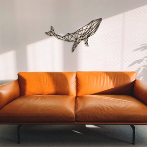 May include: A brown leather sofa with a geometric whale wall sculpture hanging above it. The sofa is in a room with white walls and a window letting in natural light.