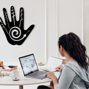 May include: A black metal wall decor in the shape of a hand with various abstract designs, including a spiral, on a white wall. A person is sitting at a table with a laptop and a book.