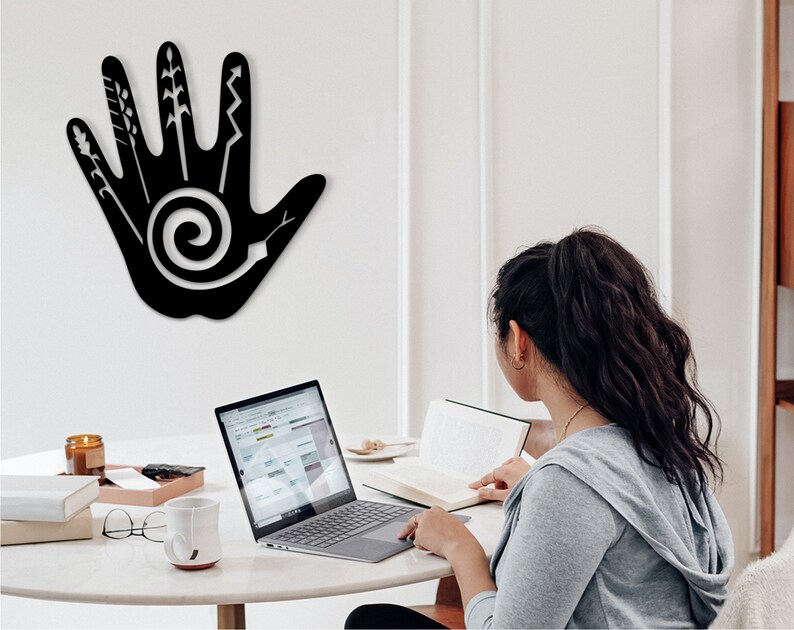 May include: A black metal wall decor in the shape of a hand with various abstract designs, including a spiral, on a white wall. A person is sitting at a table with a laptop and a book.
