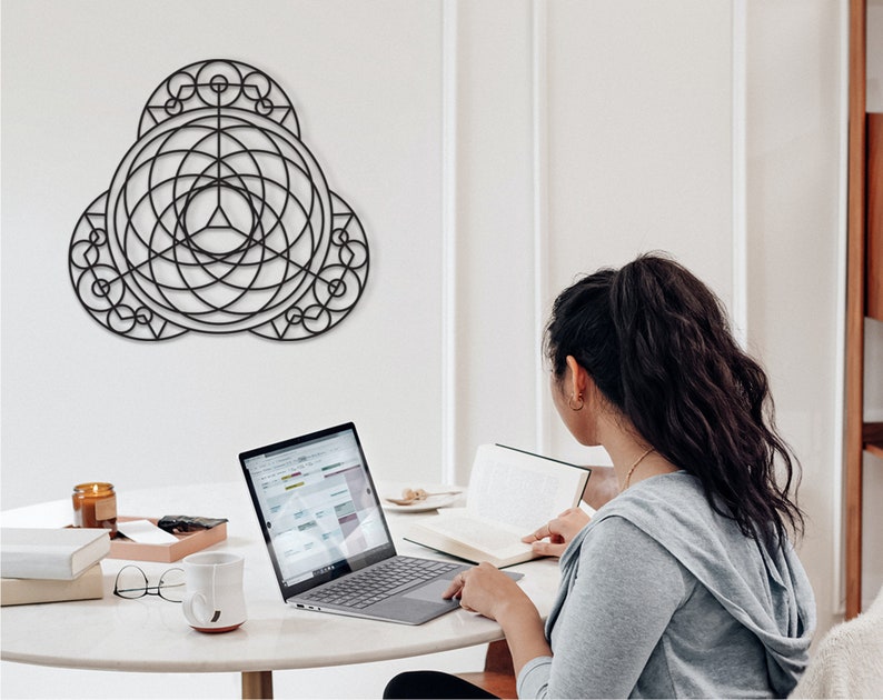 Puede incluir: Una mujer se sienta en una mesa con un port&aacute;til, un libro y una taza. Una pieza de arte geom&eacute;trico de pared cuelga en la pared detr&aacute;s de ella.