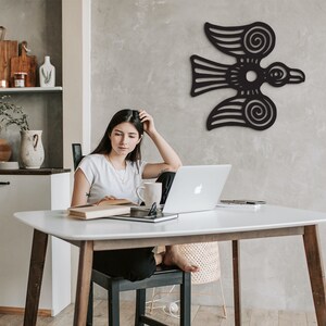 May include: A woman sits at a white table with a laptop, a book, and a mug. A black stylized bird design is on the wall behind her.