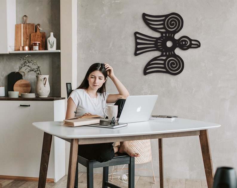 May include: A woman sits at a white table with a laptop, a book, and a mug. A black stylized bird design is on the wall behind her.
