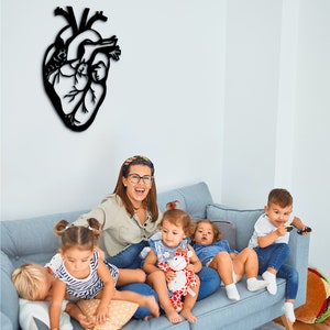 Op de afbeelding: Een groep kinderen en een volwassene zitten op een blauwe bank. De volwassene draagt een lichtblauw shirt en een bril. De kinderen dragen verschillende kleding. Er is een zwarte hartvormige wanddecoratie op de muur achter de bank.