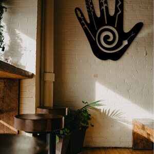May include: A black metal wall art piece in the shape of a hand with a spiral design in the palm. The hand is decorated with various lines and shapes, resembling stylized plants or flowers. The art is mounted on a white brick wall.