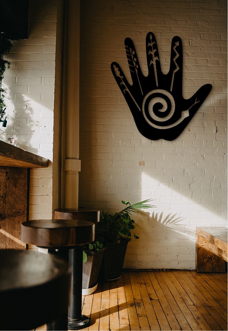 May include: A black metal wall art piece in the shape of a hand with a spiral design in the palm. The hand is decorated with various lines and shapes, resembling stylized plants or flowers. The art is mounted on a white brick wall.