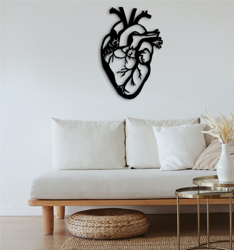 Op de afbeelding: Zwarte metalen anatomische hartwanddecoratie hangend boven een witte bank met kussens en twee gouden bijzettafels.
