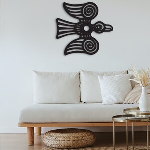 May include: Black metal wall art depicting a stylized bird with intricate details. The bird is flying with its wings spread wide. The art is mounted on a white wall above a white couch with pillows.
