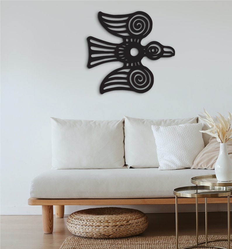 May include: Black metal wall art depicting a stylized bird with intricate details. The bird is flying with its wings spread wide. The art is mounted on a white wall above a white couch with pillows.