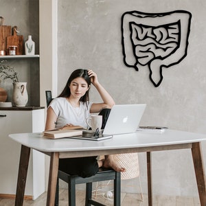May include: A woman sits at a white table with a laptop, a book, and a mug. The wall behind her features a black silhouette of the human digestive system.