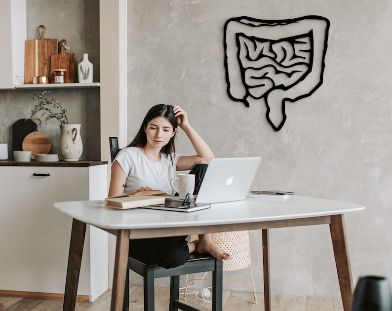 May include: A woman sits at a white table with a laptop, a book, and a mug. The wall behind her features a black silhouette of the human digestive system.