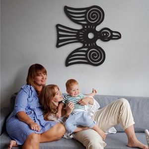 May include: Two adults and a child sit on a gray couch in front of a black wall decor of a stylized bird with intricate details. The adults are wearing striped shirts and the child is wearing a striped shirt and blue jeans.