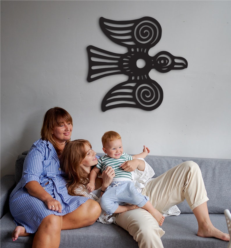 May include: Two adults and a child sit on a gray couch in front of a black wall decor of a stylized bird with intricate details. The adults are wearing striped shirts and the child is wearing a striped shirt and blue jeans.