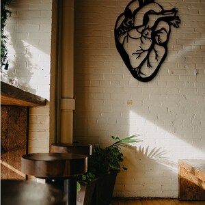 Op de afbeelding: Een zwarte metalen anatomische hartwanddecoratie die aan een witte bakstenen muur hangt. Het hart is gedetailleerd met aderen en slagaders. Het hart is in moderne stijl en zou een geweldige aanvulling zijn op elk interieur.