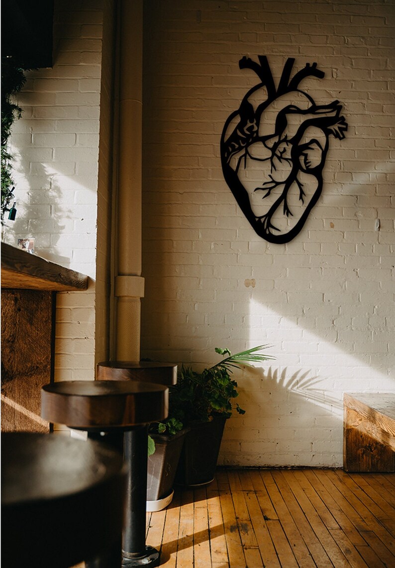 Op de afbeelding: Een zwarte metalen anatomische hartwanddecoratie die aan een witte bakstenen muur hangt. Het hart is gedetailleerd met aderen en slagaders. Het hart is in moderne stijl en zou een geweldige aanvulling zijn op elk interieur.