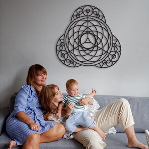 Puede incluir: Dos adultos y un ni&ntilde;o est&aacute;n sentados en un sof&aacute; gris frente a una pieza de arte de pared geom&eacute;trica negra. Los adultos est&aacute;n sonriendo y el ni&ntilde;o est&aacute; riendo. Los adultos visten ropa casual y el ni&ntilde;o lleva una camisa a rayas verde y blanca y pantalones vaqueros azules.