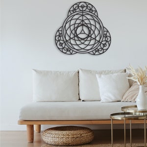 Puede incluir: Una decoraci&oacute;n de pared de metal negro con un dise&ntilde;o geom&eacute;trico, que presenta c&iacute;rculos y l&iacute;neas superpuestos. La decoraci&oacute;n est&aacute; colgada en una pared blanca sobre un sof&aacute; blanco con almohadas.