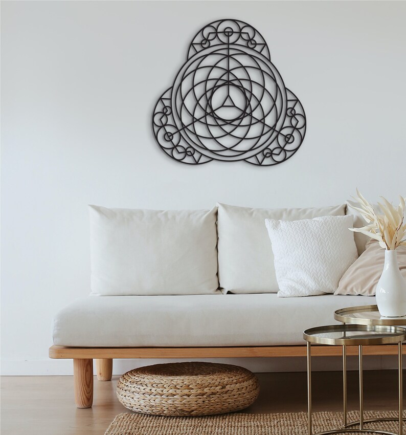 Puede incluir: Una decoraci&oacute;n de pared de metal negro con un dise&ntilde;o geom&eacute;trico, que presenta c&iacute;rculos y l&iacute;neas superpuestos. La decoraci&oacute;n est&aacute; colgada en una pared blanca sobre un sof&aacute; blanco con almohadas.