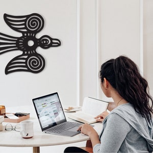 May include: A woman sits at a table with a laptop, a book, and a mug. A black stylized bird design is on the wall behind her.