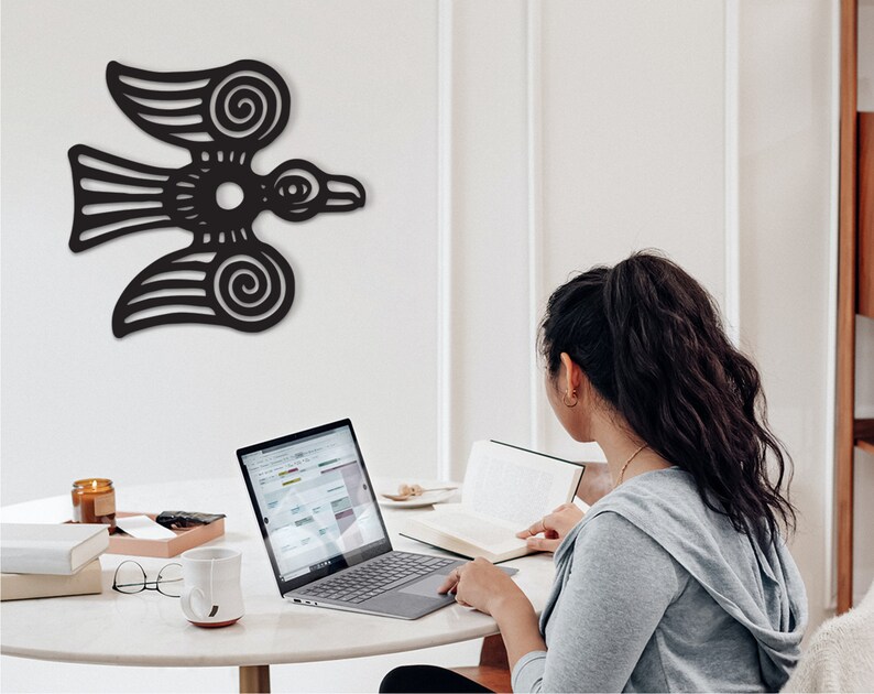 May include: A woman sits at a table with a laptop, a book, and a mug. A black stylized bird design is on the wall behind her.