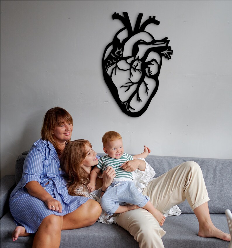 Op de afbeelding: Twee volwassenen en een kind zitten op een grijze bank. Het kind zit op de schoot van een van de volwassenen. De volwassene draagt een wit shirt en een beige broek. De andere volwassene draagt een blauw-wit gestreept shirt en een blauwe jurk. Een zwart silhouet van een hart is op de muur achter hen.