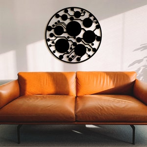 May include: A black metal wall decoration in the shape of a circle with a geometric design of lines and circles. The decoration is hanging on a white wall above a brown leather sofa.