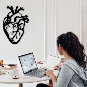 Op de afbeelding: Een zwarte metalen anatomische hartwanddecoratie die aan een witte muur hangt. Een persoon zit aan een tafel met een laptop en een boek.