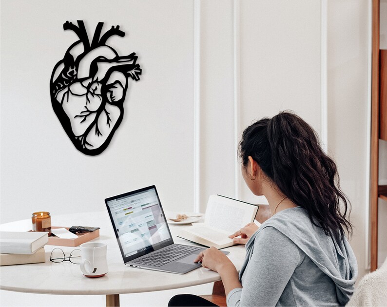 Op de afbeelding: Een zwarte metalen anatomische hartwanddecoratie die aan een witte muur hangt. Een persoon zit aan een tafel met een laptop en een boek.