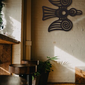 May include: A black metal wall art piece depicting a stylized bird with intricate details. The bird is flying with its wings spread wide and has a circular design in the center of its body. The art piece is mounted on a white brick wall.